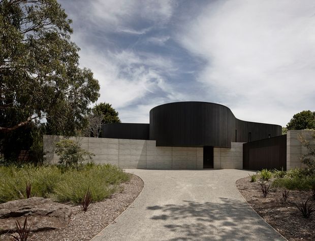 Street-facing view of a sculptural contemporary residence with a dark, curved upper facade set behind pale horizontal concrete boundary walls. A broad textured concrete driveway leads to a recessed dark entry, surrounded by restrained native-style landscaping, gravel beds, grasses, rocks, and mature trees.