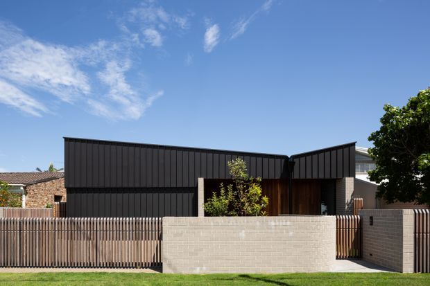 Street-facing exterior of a contemporary single-storey residence with dark vertical metal cladding, contrasting pale brick masonry, timber-toned entry detailing, and a low masonry front wall with vertical slatted fencing. The composition features angular pitched roof forms and a clean, minimalist facade under a bright blue sky.