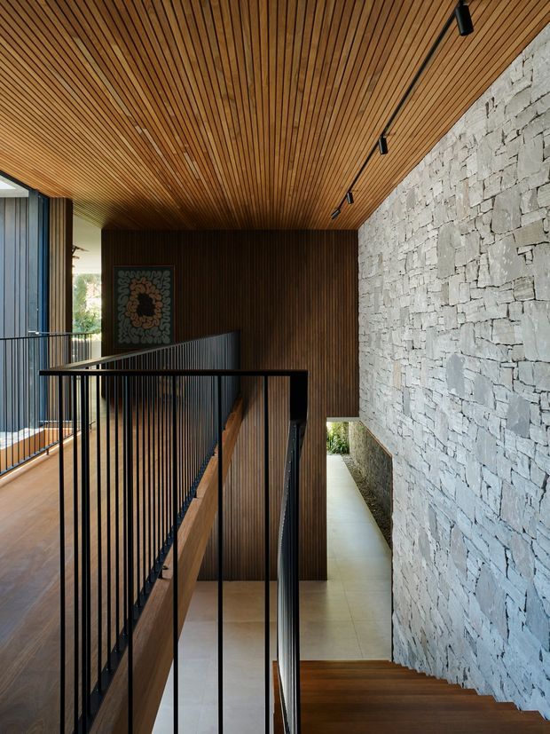 A modern interior stairway and landing with a warm slatted timber ceiling and matching dark timber wall panelling. A large rough-faced pale stone wall runs alongside the stairs, contrasting with slim black metal balustrades, timber stair treads, and a light tiled floor. Track lighting is integrated into the ceiling, and a glazed opening brings in daylight.