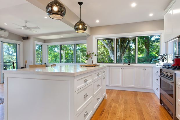 Bright, spacious white kitchen with a large central island, extensive shaker-style cabinetry, pale benchtops, stainless-steel appliances, and warm timber flooring. Large windows overlook leafy trees, while pendant lights and recessed lighting illuminate the open room.