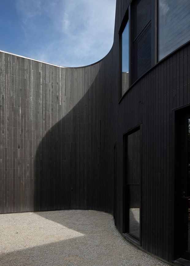 A stark, sculptural exterior courtyard enclosed by tall, curved walls clad in narrow vertical dark timber boards. Black-framed glazing and doors line the right-hand building facade, while pale gravel covers the ground. Strong sunlight creates dramatic geometric shadows across the dark facade, with a small portion of blue sky visible above.