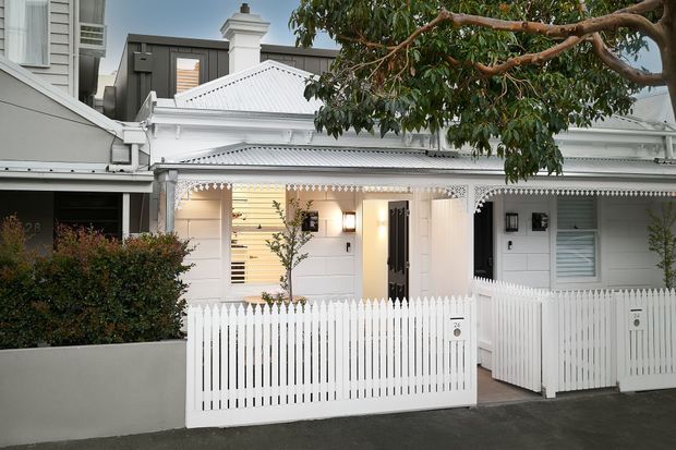 Street-facing view of a compact white heritage-style townhouse or cottage facade, featuring ornate decorative fretwork, a corrugated metal pitched roof, white picket fencing, timber weatherboard cladding, and a small covered entry porch. A darker contemporary building is visible behind it.
