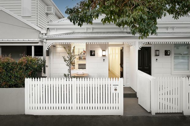 Front exterior of a white weatherboard residence with ornate fretwork trim, a covered entry porch, white picket fencing, and a small landscaped front garden. The facade features traditional detailing, a dark front door, horizontal cladding, and decorative eaves.