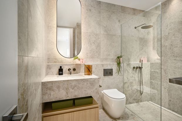 Contemporary bathroom with large-format grey stone-look wall and floor tiles, a floating stone vanity with integrated basin, rounded vertical mirror, wall-mounted toilet, and frameless glass walk-in shower with overhead rain shower and handheld fixture. Warm timber and green accents add contrast to the neutral finishes.