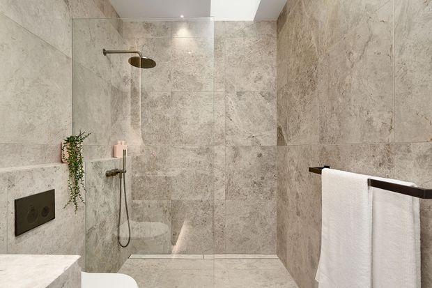 Contemporary walk-in bathroom shower with large-format beige-grey stone-look tiles covering the walls and floor. A frameless glass shower screen encloses a rain shower and handheld fixture, with dark metal fittings, a wall-mounted flush plate, recessed ceiling lighting, a narrow skylight or clerestory opening, and a wall-mounted towel rail with white towels. A small trailing plant adds greenery.