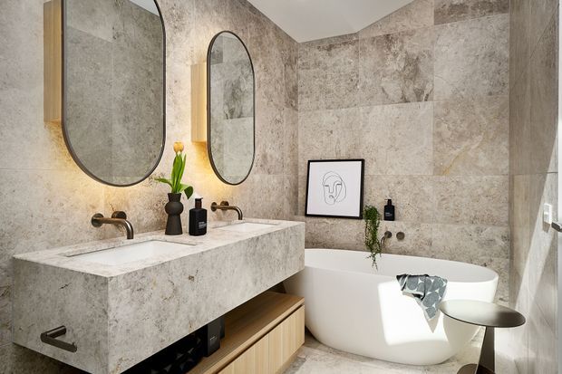 A refined contemporary bathroom featuring a large double vanity clad in pale veined stone, two wall-mounted brushed-metal faucets, arched black-framed mirrors, full-height stone-look wall tiles, a freestanding white bathtub, and warm timber storage below the vanity. Black accessories, greenery, and framed artwork provide restrained accents.
