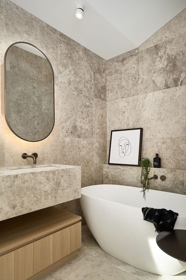 A refined contemporary bathroom featuring a freestanding white bathtub, wall-mounted aged-bronze tapware, a stone-clad floating vanity with timber cabinetry, and a large rounded-rectangle mirror. The walls and floor are finished in richly veined beige-grey stone tiles, with soft integrated lighting and restrained artwork and greenery.