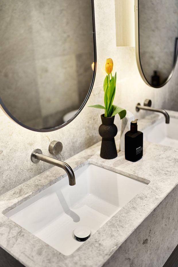 Close-up of a contemporary bathroom vanity featuring a white undermount basin set into a pale grey marbled stone benchtop, brushed metal wall-mounted tapware, grey textured wall finish, and large rounded mirrors. Black soap dispensers and a small vase with a yellow tulip add contrast.