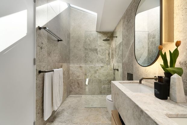 Bright contemporary bathroom featuring full-height grey-beige stone-look wall and floor tiles, a frameless glass shower with ceiling skylight, wall-mounted rainfall shower, white toilet, and a substantial stone vanity with integrated basin. A rounded black-framed mirror and warm timber cabinetry add contrast.