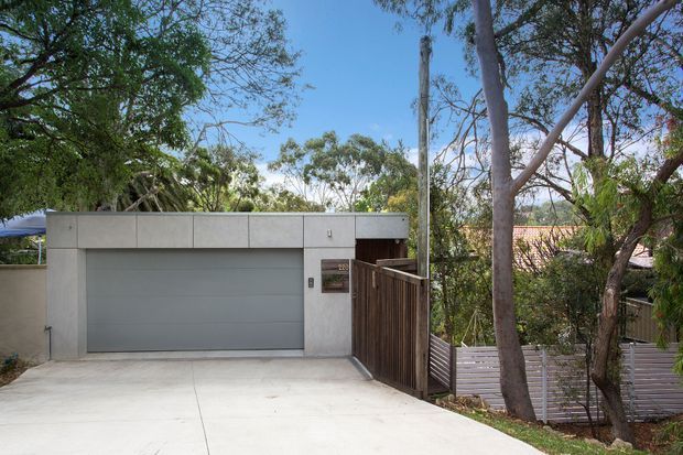 Street-facing view of a compact modern residential garage and entry facade. The building has a flat roof, smooth light-grey rendered or concrete walls, a wide sectional garage door, a dark timber pedestrian gate, and a pale concrete driveway. Mature trees and timber screening surround the property, with a tiled roof of the residence partially visible behind.