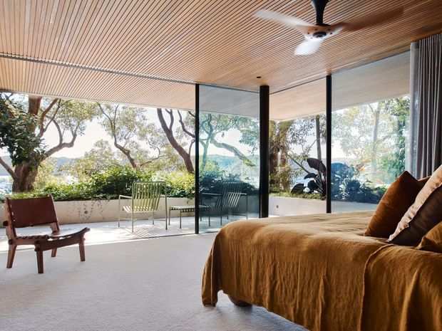 A bright, spacious bedroom with floor-to-ceiling glazing opening toward a planted outdoor terrace. The room features a broad low-profile bed dressed in warm brown linen, a timber slatted ceiling, dark slender structural window frames, pale carpet, and a sculptural ceiling fan. Outdoor chairs and established trees are visible beyond the glazing.