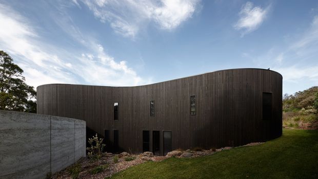 Exterior view of a sculptural, curved single-storey building set into a landscaped sloping site. The facade is clad in dark vertical timber boards with narrow windows and doors, alongside a raw concrete boundary or retaining wall. The rounded roofline and organic form contrast with the grass and planted surroundings under a bright sky.