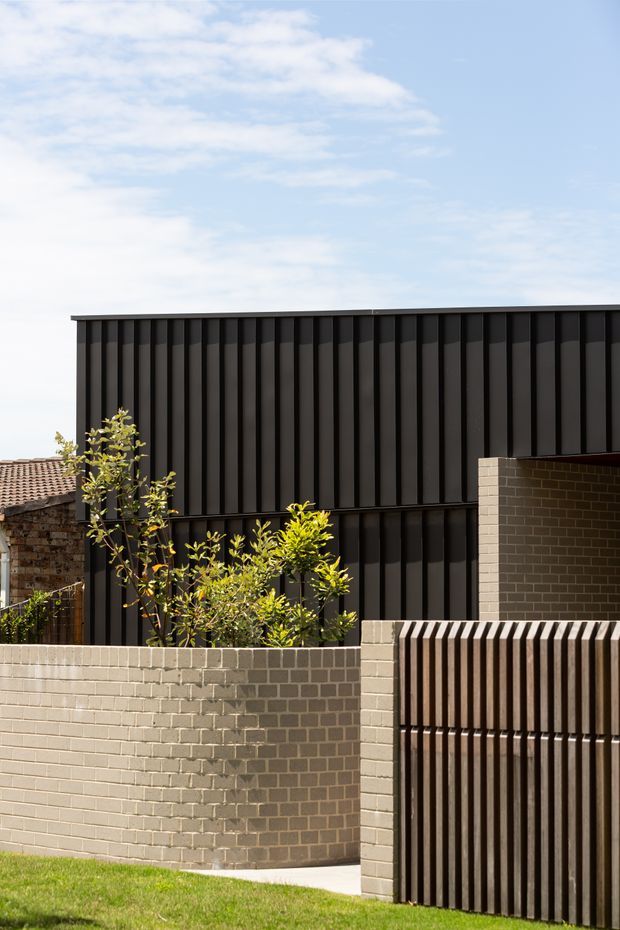 Exterior architectural photograph of a contemporary residential facade. A tall black vertically ribbed metal-clad wall forms the main backdrop, with pale grey-beige brick boundary walls and a matching brick garage or entry volume. A dark vertical slatted gate occupies the foreground, with sparse landscaped planting and lawn at the base under a bright partly cloudy sky.