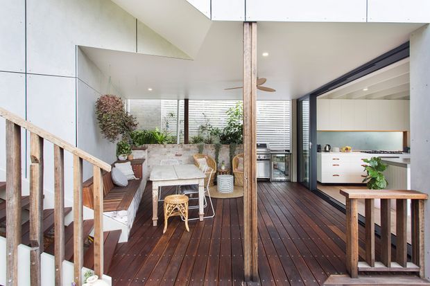 Bright contemporary open-plan living and kitchen area opening onto a timber deck. The space features dark timber decking, white walls and cabinetry, extensive louvred glazing, exposed timber posts, a compact dining setting, built-in bench seating, and a modern white kitchen with a timber benchtop. A stair railing is visible in the foreground.