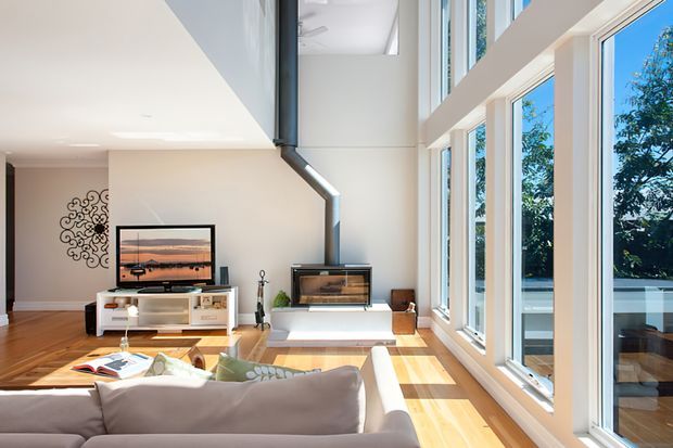Bright contemporary living room with a large upholstered sectional sofa facing a television and a modern fireplace. The room features warm timber flooring, white walls and built-in joinery, dramatic floor-to-ceiling glazing, and a double-height volume with abundant natural light. A leafy exterior is visible through the windows.