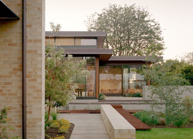 Exterior view of a contemporary, low-profile residential building surrounded by landscaped gardens. The composition shows warm pale brick walls, broad horizontal roof planes, extensive floor-to-ceiling glazing, dark metal framing, and a timber boardwalk leading toward the glazed living spaces. Layered garden beds and a low masonry wall create a calm courtyard-like approach.