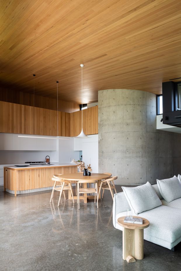 Open-plan contemporary interior combining a kitchen, dining area and living room. The space features extensive warm timber lining across the ceiling and kitchen cabinetry, a polished concrete floor, white lower cabinetry and walls, a curved exposed concrete feature wall, black-framed glazing, a round timber dining table with light timber chairs, and a pale upholstered sofa. Minimal pendant lights and restrained furnishings create a warm, refined, modern atmosphere.