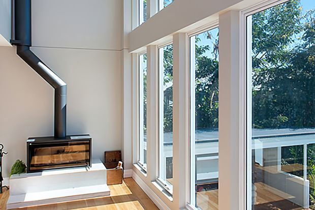 Bright contemporary living room with a freestanding wood-burning fireplace, white built-in hearth, timber flooring, and extensive floor-to-ceiling glazing overlooking greenery and an exterior deck or balcony. The room has a high ceiling and strong natural light.