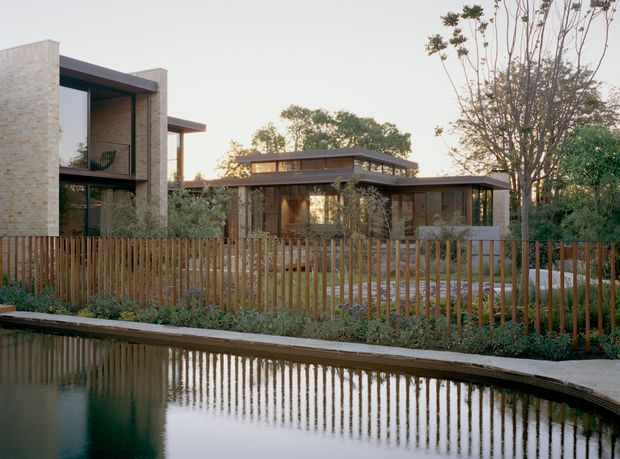 Exterior view of a contemporary low-rise residence beside a reflective water feature. The home combines pale masonry walls, extensive dark-framed glazing, flat projecting roof planes and warm timber detailing. A vertical timber privacy fence and landscaped planting run along the water's edge, with mature trees surrounding the property.