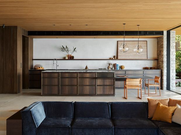 Open-plan interior featuring a dark timber kitchen with a large island, integrated cabinetry, stone or light-toned benchtops, and a long open shelf against a pale wall. A low navy sectional sofa occupies the foreground, with dining-style timber chairs beside the island. The ceiling is clad in linear timber boards, while full-height glazing at right opens toward greenery. Pendant globe lights, artwork, and restrained styling create a warm, refined contemporary atmosphere.