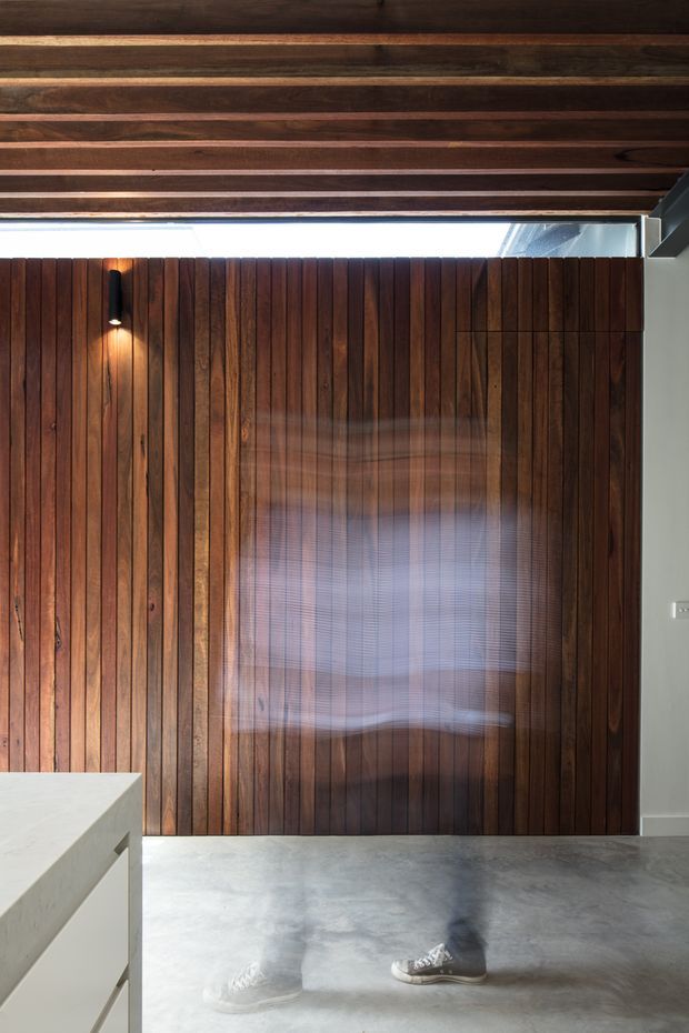 Interior view featuring a full-height dark timber-lined feature wall with a concealed or glazed opening, exposed timber ceiling beams, polished grey concrete flooring, and a white built-in island or cabinet in the foreground. A slim clerestory-like glazed strip brings in daylight, while a wall-mounted cylindrical light provides accent illumination. The image has a raw, contemporary architectural character; a faint human reflection or motion blur is visible on the timber wall and floor.