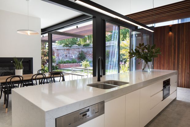 Contemporary open-plan kitchen and dining area centered on a large white island with integrated sink, dishwasher, drawers and built-in oven. Black-framed floor-to-ceiling glazing opens toward a landscaped courtyard, while a dark timber slatted ceiling and vertical timber wall create warmth and contrast. A dining table, pendant light and indoor plant are visible.