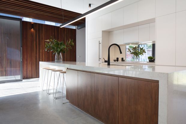Contemporary kitchen with a large white stone island and integrated breakfast bar, dark timber cabinetry, white full-height cabinetry, and a black gooseneck faucet. The space features polished concrete flooring, a timber slatted ceiling, and a richly toned vertical timber wall, creating a refined contrast between warm wood and crisp white surfaces.