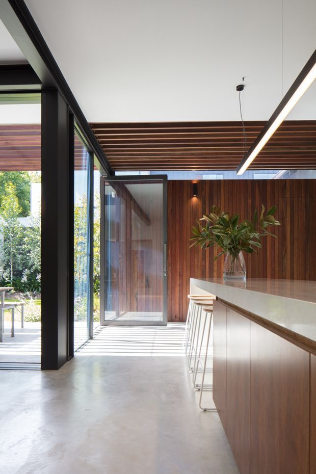 A bright, contemporary kitchen interior with a long timber-clad island and pale benchtop, slim bar stools, full-height black-framed glazing, and a partially open glass door. The ceiling features exposed timber battens and a linear pendant light, while a planted arrangement adds greenery. Polished concrete flooring and vertical timber wall lining create a warm natural-industrial feel.