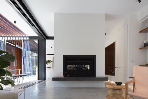Bright contemporary living room with a minimalist white fireplace wall, black built-in fireplace insert and dark hearth. Full-height black-framed glazing opens toward a covered outdoor area with timber slatted screening. The room includes pale polished flooring, concealed or flush white cabinetry, timber shelving, and sculptural timber furniture.