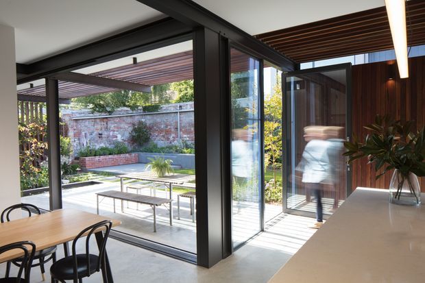 Contemporary open-plan kitchen and dining area opening directly onto a landscaped courtyard through extensive black-framed glazed doors. The space features a pale concrete floor, a large light-toned kitchen island or benchtop, dark timber wall panelling, exposed dark ceiling battens, and a timber dining table with black chairs. A brick boundary wall, pergola, paved terrace, and outdoor dining furniture are visible beyond the glazing.