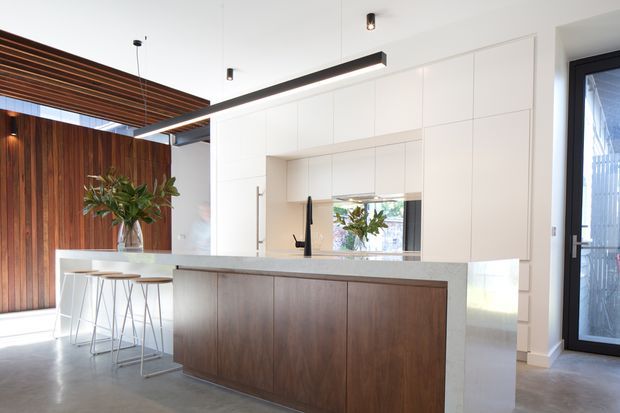 Bright contemporary kitchen with a large island featuring dark timber cabinetry and a pale stone benchtop. White full-height built-in cabinetry lines the rear wall, with an integrated sink and black tap, while a timber slatted ceiling feature and vertical timber wall add warmth. The space has polished concrete flooring, black-framed glazing, minimalist linear pendant lighting, and a small island dining area.