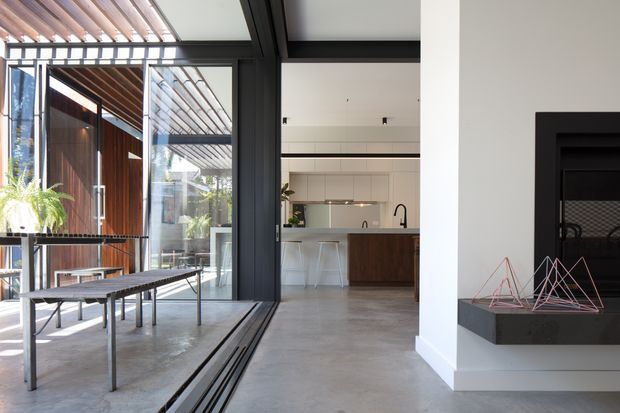 Bright contemporary open-plan interior with polished concrete flooring, a minimalist kitchen visible beyond large black-framed glazed doors, and a recessed dark fireplace on the right. The space opens to a covered outdoor terrace with timber screening, pergola-like roof structure, and a long bench. Strong indoor-outdoor connection, clean white walls, dark framing, and warm timber accents define the design.