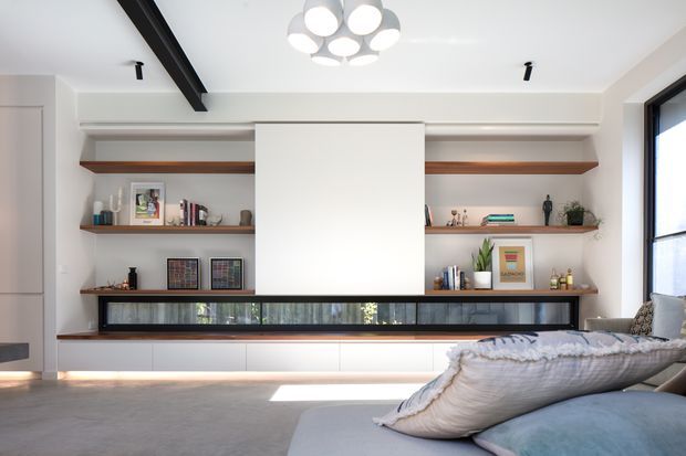 Bright contemporary living room with a low built-in media wall and extensive open timber shelving, white cabinetry, and a dark-framed horizontal window or display opening. A large pale sectional sofa occupies the foreground, while a sculptural globe pendant hangs from the white ceiling. A dark exposed ceiling beam adds contrast, and daylight enters through the tall side window.