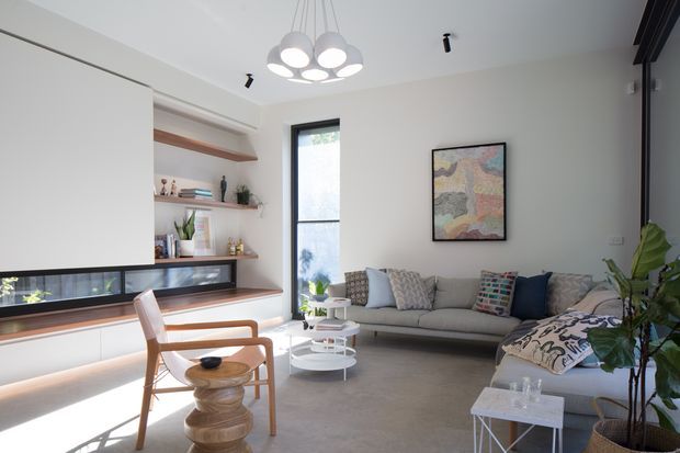 Bright, contemporary living room with a neutral palette, polished concrete flooring, a long low built-in media/storage wall with open timber shelving, a grey sofa, sculptural white coffee tables, a timber lounge chair, indoor plants, and a modern multi-globe pendant light. Tall glazing brings in abundant daylight, and a colourful framed artwork provides a focal accent.