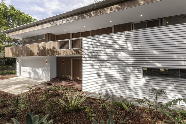 A contemporary two-storey residential facade set within landscaped grounds. The house combines white horizontal weatherboard cladding, warm vertical timber wall lining, a broad cantilevered upper-level balcony, dark-framed windows, recessed exterior downlights, and an integrated white garage. Low-maintenance planting occupies the foreground, while mature trees cast dappled shadows across the facade.