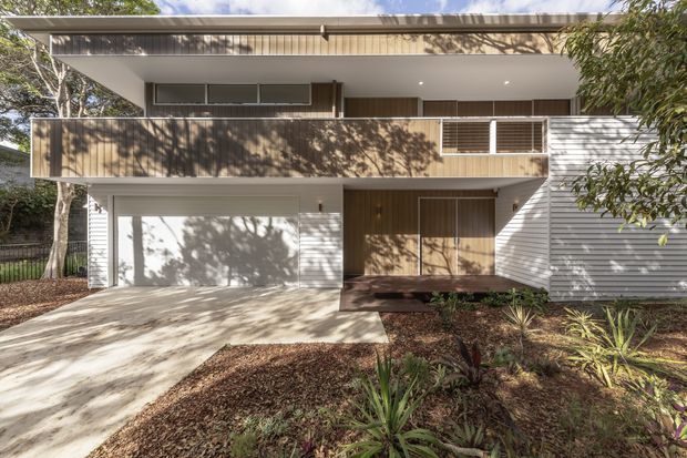 Exterior view of a contemporary two-storey residence set among established trees and landscaped garden beds. The house features a broad flat roof, projecting upper-level balcony or terrace with a solid brown privacy screen, white horizontal weatherboard-style cladding, timber-look vertical wall cladding around the entry, and an integrated garage. A concrete driveway and walkway lead to the recessed front entrance.