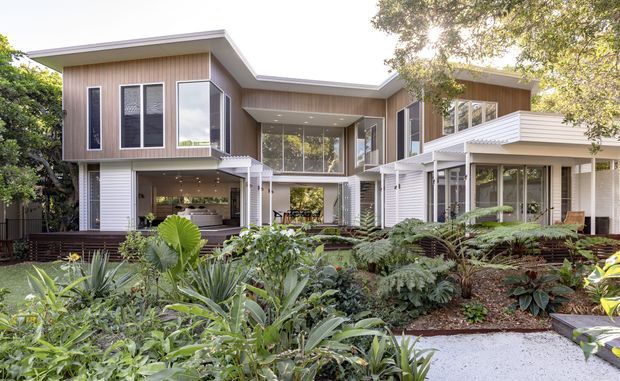 A contemporary multi-wing residential house viewed from a landscaped garden. The façade combines light timber-look vertical cladding, white horizontal cladding, extensive glazing, projecting roof planes, and covered outdoor living areas around a central glazed connection. Dense tropical planting and a pale gravel path occupy the foreground.