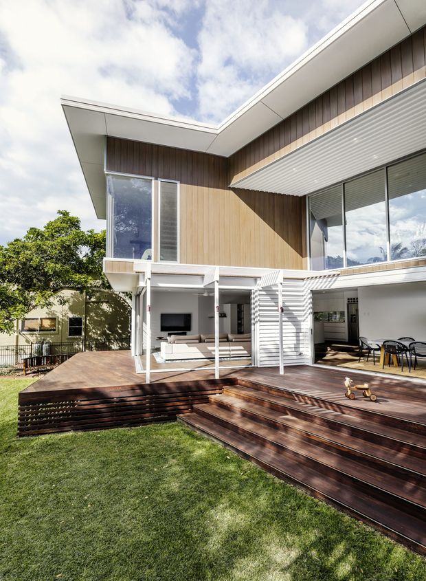 Exterior rear view of a contemporary two-storey home with a light timber-clad upper level, broad white roof overhangs, extensive glazing, external roller shades, and a covered outdoor living area. A large dark timber deck with tiered steps extends into the lawn, with a secondary paved dining area and white slatted screening beneath the upper floor.