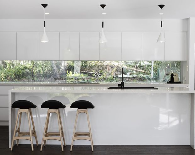 Bright contemporary kitchen with a large white island and integrated seating for three. The room features extensive white flat-panel cabinetry, a pale stone benchtop, dark timber flooring, a black undermount-style sink and tap, and three sculptural pendant lights. A long horizontal window provides views to planted greenery outside.
