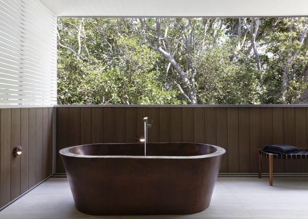 A refined, semi-open bathroom with a freestanding dark bronze or brown bathtub positioned centrally beneath a wall-mounted bath filler. Dark timber wall panelling wraps the lower walls, while a white horizontal slatted screen and large open glazing provide privacy, filtered daylight, and views into dense greenery. Pale large-format floor tiles, a small stool, and restrained contemporary detailing create a calm, luxurious indoor-outdoor bathing space.