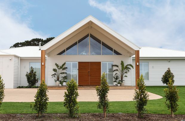 Front exterior of a single-storey white weatherboard house with a prominent gabled entry porch, exposed timber posts, wide triangular glazing, a warm timber double front door, and symmetrical landscaping with clipped shrubs and lawn.