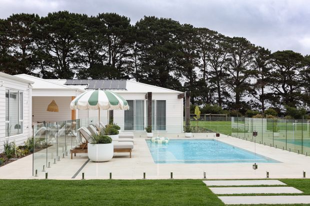 A bright, contemporary single-storey white-clad residence opens onto a paved pool terrace. The outdoor area includes a rectangular swimming pool enclosed by frameless glass fencing, a covered patio with pale outdoor seating, a striped umbrella, large planters, and neatly arranged lawn and stepping stones. Tall mature trees form the background, and solar panels are visible on the low roof.