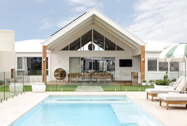 Bright rear exterior view of a coastal-style single-storey residence with a prominent gabled roof, white horizontal weatherboard cladding, timber posts, and a glazed triangular gable. The house opens onto a lawn and paved pool terrace with a rectangular swimming pool, outdoor seating, and a striped parasol.