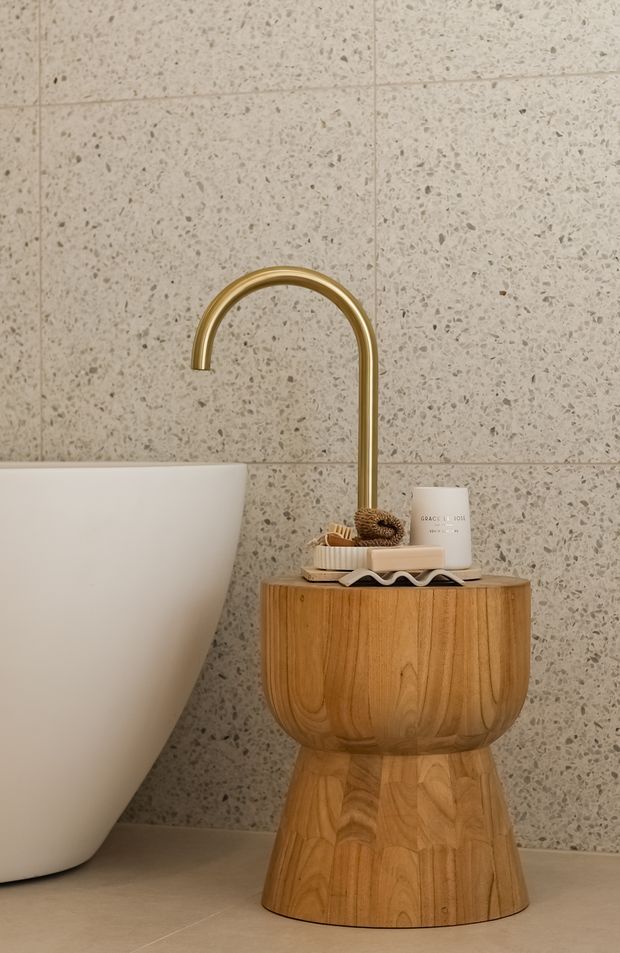 Close-up of a refined bathroom vignette featuring a freestanding white bathtub, a tall curved brass/gold tap, and a sculptural natural timber pedestal side table with toiletries. Large-format speckled stone-look wall tiles and a warm neutral tiled floor create a calm, luxurious setting.