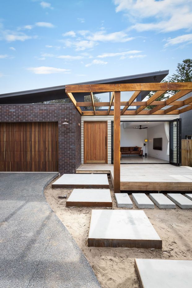 Exterior view of a contemporary single-storey residence with a dramatic skillion roof, dark brick garage wall, timber-clad garage door, and a timber-framed covered outdoor deck. Large bi-fold doors open into a living room, while stepping slabs cross a sandy landscaped foreground.