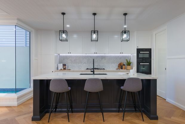 Bright, refined contemporary kitchen with a large dark island and three upholstered bar stools, white perimeter cabinetry, a textured full-height splashback, integrated black appliances, pendant lighting, and light herringbone timber flooring. A glazed pool or outdoor area is visible at the left.