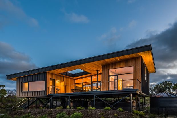 Exterior dusk photograph of a contemporary elevated home with a broad single-slope roof, dark metal cladding, warm timber-lined walls and soffits, extensive glazing, and a raised structural frame above the landscaped ground. A glazed roof opening/skylight and sheltered outdoor deck are visible.