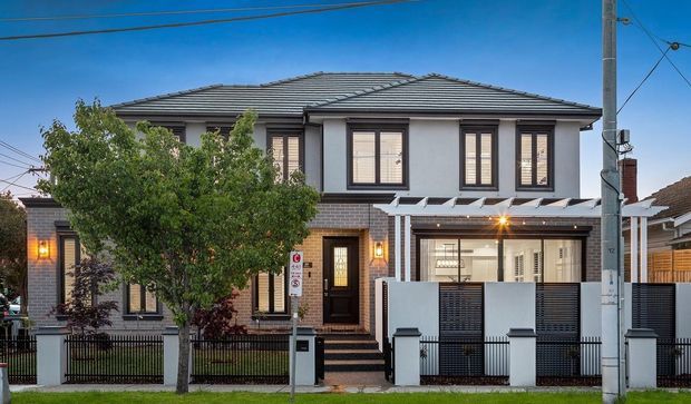 Street-facing view of a contemporary two-storey attached townhouse with a hipped tiled roof, light rendered upper facade, dark brick lower facade, tall windows, and a fenced front entry courtyard. Utility poles and neighbouring homes are visible in the suburban streetscape.