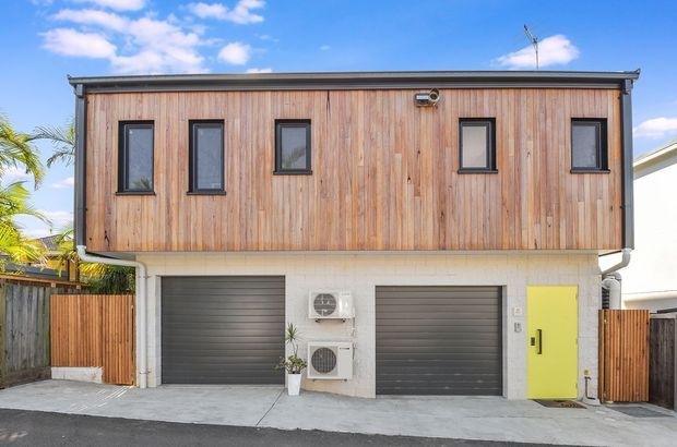 Exterior view of a compact multi-storey urban building with a distinctive upper level clad in vertical natural timber boards. The lower level is finished in light masonry or painted blockwork and contains two dark grey garage doors, a bright lime-green pedestrian door, and visible external air-conditioning units. Narrow vertical windows with dark frames punctuate the timber facade. Timber privacy fencing and a concrete driveway are visible, with a blue sky and palms in the background.