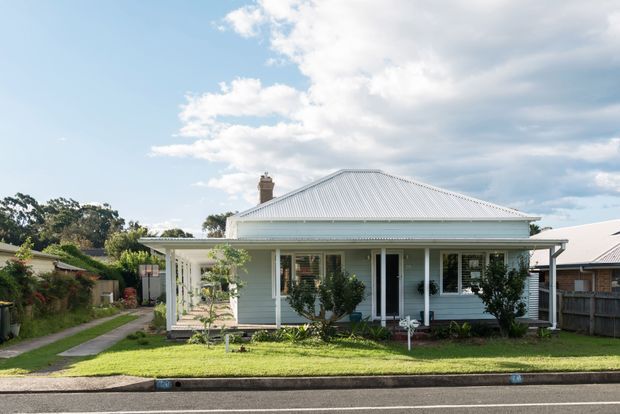 Front exterior view of a single-storey detached cottage-style home with a pale weatherboard facade, broad corrugated metal hipped roof, covered front verandah, white posts and trim, and a modest landscaped front garden. The house sits in a suburban streetscape with neighbouring homes visible at the sides.